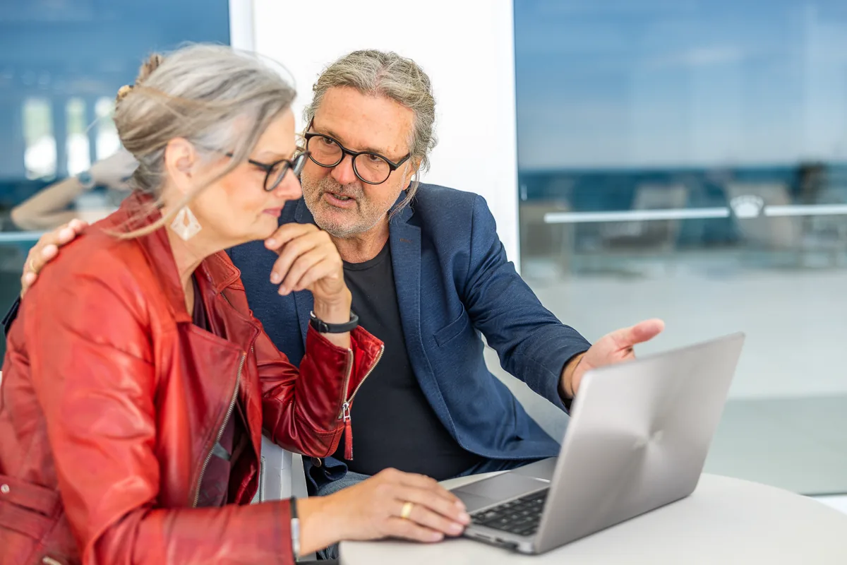 bruno-dagmar-laptop-terrasse Eine Frau in roter Lederjacke und ein Mann im blauen Sakko sitzen an einem Tisch auf einer Terrasse am Meer und sehen gemeinsam auf einen aufgeklappten Laptop.