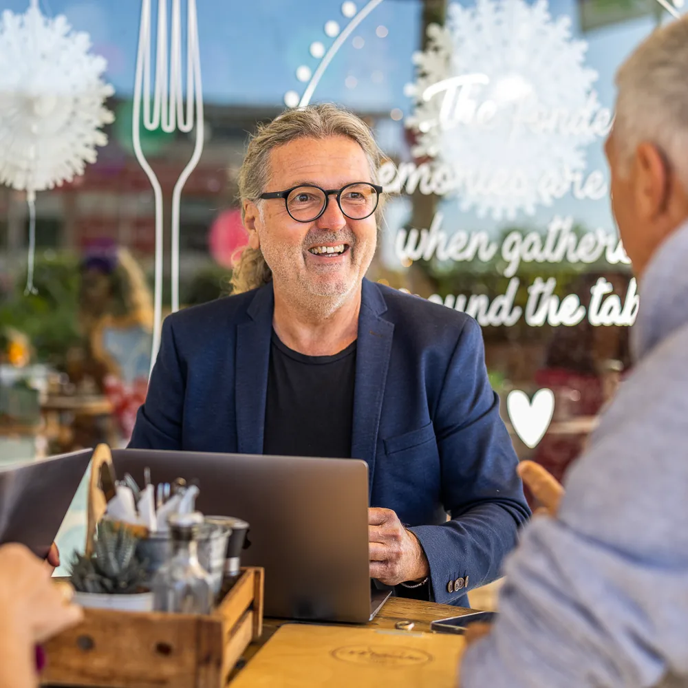 Ein Mann mit Brille und blauem Sakko sitzt lachend an einem Cafétisch hinter einem aufgeklappten Laptop, im unscharfen Vordergrund sein Gegenüber.