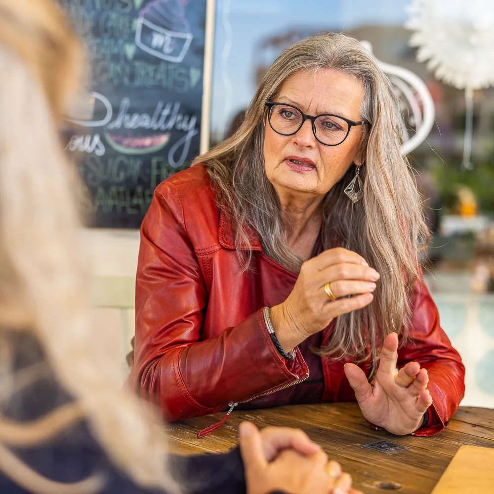Eine Frau mit langen grauen Haaren, Brille und roter Lederjacke sitzt an einem Cafétisch und erklärt ihrem Gegenüber etwas mit den Händen.